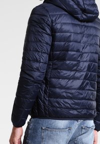 Marinblå pufferjacka med huva, med en vadderad design och glänsande, lätt material, visad bakifrån med synliga stygn.