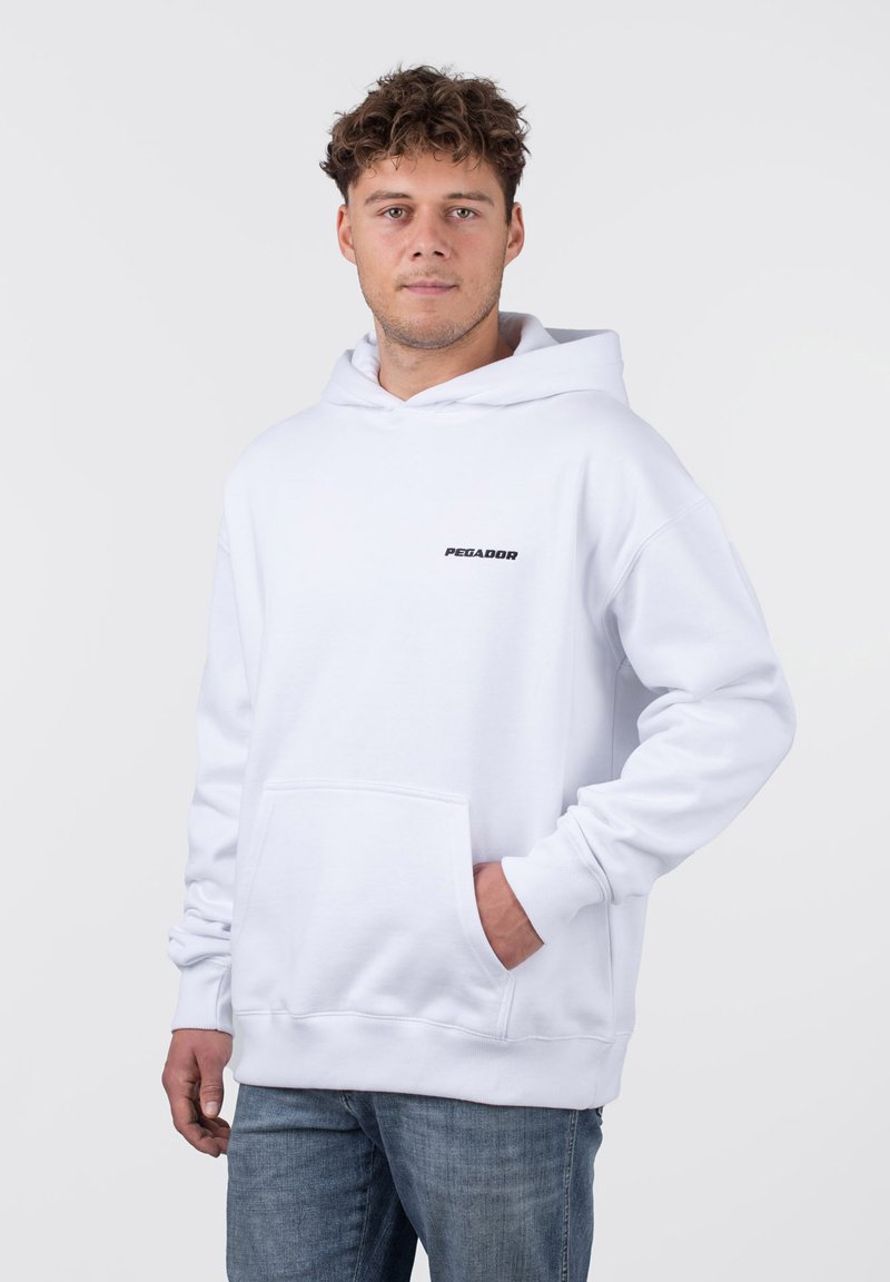 Weißer Hoodie aus softem Stoff mit einer Fronttasche, Kapuze mit Kordelzug und einem schwarzen "PEGADOR"-Logo auf der Brust. Getragen mit blauen Jeans.