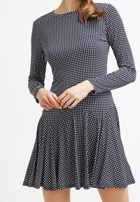 Kvinna i en svartvit rutmönstrad långärmad klänning med figurnära midja och utsvängd kjol, stående mot en enfärgad bakgrund.