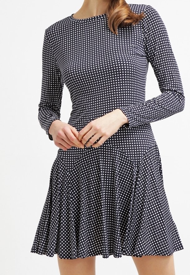 Kvinna i en svartvit rutmönstrad långärmad klänning med figurnära midja och utsvängd kjol, stående mot en enfärgad bakgrund.