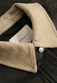 Gros plan sur un col en velours côtelé beige sur une veste sombre, bouton-pression métallique visible, avec une étiquette portant la mention "NOELLA" à l’intérieur du col.