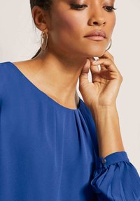 Blaue Bluse mit weitem Halsausschnitt, gefältelten Details und gerafften Ärmeln. Glattes, leichtes Material mit metallischem Akzent am Bündchen.