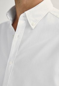 Camicia bianca in cotone con bottoni. Presenta un colletto strutturato, una texture sottile e bottoni perla. Vista ravvicinata del colletto.