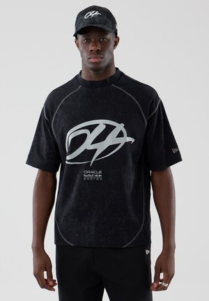 Jeune homme portant un t-shirt noir Oracle Red Bull Racing et une casquette assortie, debout devant un fond gris uni.