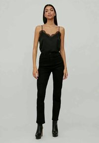 Camisola nera in satin con pizzo, abbinata a jeans neri a vita alta e stivali neri alla caviglia. Il modello è in piedi con le mani lungo i fianchi.