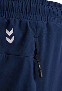 Granatowe sportowe spodenki z elastycznym pasem, kieszenią na zamek i białymi akcentami w kształcie szewronu po boku. Miękki, lekki materiał.