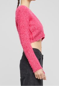 Kvinna iklädd en ljusrosa fluffig kortärmad tröja med lång ärm och högmidjade svarta jeans, visad i profil mot en enfärgad bakgrund.