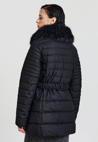 Cappotto imbottito nero con collo in ecopelliccia, vita incinturata e cuciture verticali. Il materiale ha una texture liscia e lucida.