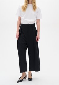 Svarta vida byxor med veck och hög midja, kombinerade med en vit t-shirt med korta ärmar och svarta spetsiga klackskor.