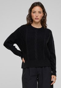 Zwarte gebreide trui met textuur kabelpatronen, een ronde halslijn en losse mouwen. Draagt een donkere broek.