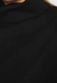 Primer plano de una persona que lleva un suéter negro de cuello alto, con el cabello largo y claro visible en el borde.