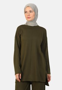 Olive green long-sleeve top in textured fabric with a round neck, side slits, and relaxed fit, paired with a light gray hijab.
