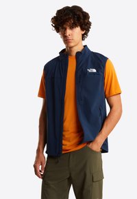 Junger Mann mit lockigem Haar, der eine marineblaue ärmellose Weste über einem orangefarbenen T-Shirt und olivgrünen Cargo-Hosen trägt, mit einer Hand in der Hosentasche stehend.