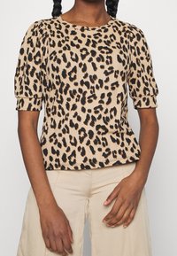 Top s leopardím vzorem vyrobený z texturované tkaniny. Má krátké nafouknuté rukávy a kulatý výstřih. Barvy zahrnují béžovou a černou.