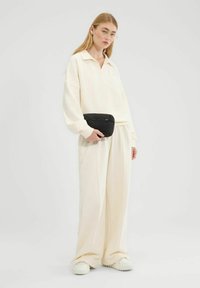 Jasny kremowy oversize'owy sweter z kołnierzykiem i pasującymi spodniami o szerokich nogawkach. Akcesoria to czarna torebka na zamek błyskawiczny i jasne obuwie.