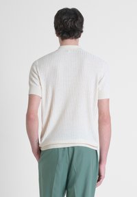 Maglione a maniche corte in crema a coste con una texture sottile e un colletto classico, abbinato a pantaloni verde chiaro, visto da dietro.