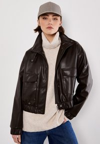 Braune Kunstleder-Shortjacke mit Druckknopfkragen, zwei Fronttaschen, elastischem Saum, getragen über einem hellbeigen Pullover und blauen Jeans.
