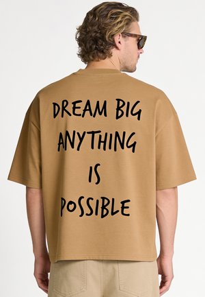 Homme aux cheveux bouclés et lunettes de soleil portant une chemise marron avec "DREAM BIG ANYTHING IS POSSIBLE" imprimé en noir dans le dos.