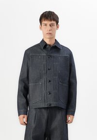 Denimjakke i mørkeblå med knaplukning, to forlommer og kontrastsyning. Har en spidse krave og lige snit.