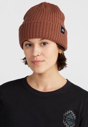 Brown ribbed knit beanie featuring a folded cuff and a small black logo patch. Worn with a black crewneck shirt.