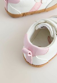 Bela otroška čevlja z roza poudarki in teksturirano gumijasto podplato. Ima jeziček z ježkom in majhno zanko za enostavno obuvanje.
