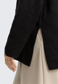 Hand rust naast een beige rokzak, met een zwarte gebreide trui met textuur en een zijsplit.