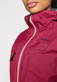 Vinrød vandtæt jakke med lyserød lynlås, sort beslag og små lommer på brystet. Har en høj krave og glat tekstur.