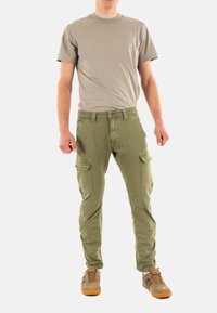 Pantaloni cargo color khaki con molte tasche a toppa, abbinati a una maglietta a girocollo grigia chiara. L'outfit include scarpe marroni con accenti scuri.