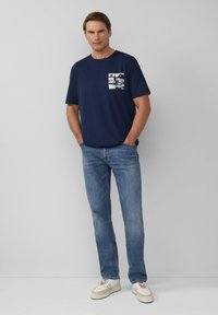 Marineblauwe katoenen t-shirt met een grafische zakprint, gecombineerd met lichtblauwe denim jeans en witte sneakers. Model staat met handen in de zakken.