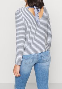 Pasiasty top z długim rękawem w kolorze szarym i białym, z wycięciem na plecach i niebieską satynową wstążką. Materiał wydaje się miękki i swobodny.