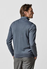 Pull en maille noir à manches longues avec un col haut, texture lisse, bords côtelés et ourlet. Coupe ajustée, porté sur un pantalon gris.