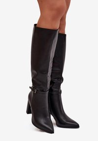 Bottes noires en cuir montant jusqu'aux genoux avec un bout pointu, un talon bloc et un détail de boucle décorative sur le côté. Texture lisse avec un design relâché.