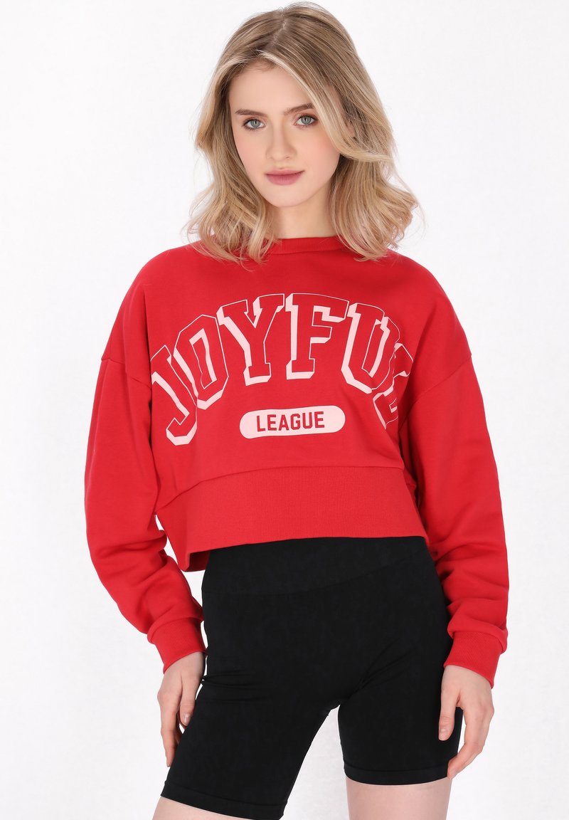 Piros rövidített pulóver, fehér és sötétkék "JOYFUL" és "LEAGUE" felirattal. Hosszú ujjú, bordás mandzsettával és laza fazonnal. Fekete biciklis sort.