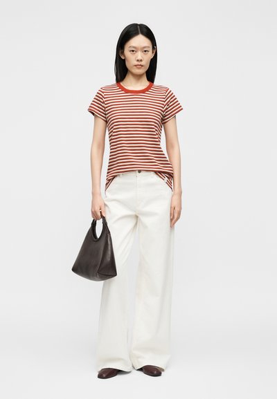 Γυναίκα στέκεται φορώντας μια κόκκινη και λευκή ριγέ μπλούζα, λευκό παντελόνι με φαρδιά μπατζάκια, κρατάει μια μαύρη τσάντα και φοράει καφέ παπούτσια.