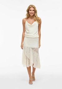 Top cami en satin blanc crémeux avec un décolleté en V, associé à une jupe en dentelle transparente avec un design en couches et des motifs texturés. Talons aiguilles argentés.