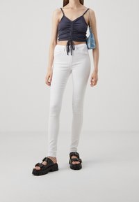 Top corto navy con detalles fruncidos, combinado con pantalones vaqueros skinny blancos. Sandalias negras de plataforma con tiras y tachuelas decorativas. Pequeño bolso azul.