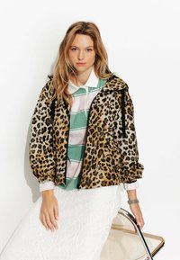 Veste en imprimé léopard avec fermeture éclair noire, poignets élastiques et capuche. Portée par-dessus un polo rayé vert et blanc et une jupe blanche texturée.