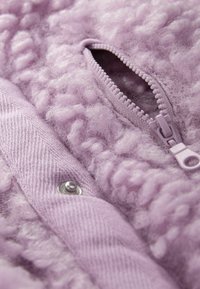 Suddig lavendelfärgad tyg med en texturerad yta, med en dragkedja och en metalldragknapp. Mjuk, fluffig textur med ett vävt mönster.
