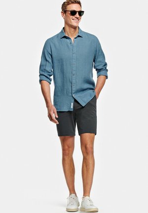 Homme souriant portant des lunettes de soleil, une chemise bleue à manches longues boutonnée avec les manches retroussées, un short noir et des baskets blanches, debout avec une main dans la poche.