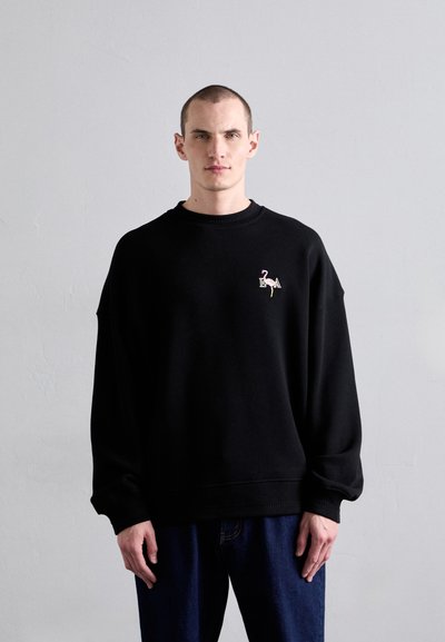 Sudadera de algodón negra con un corte relajado, cuello de canalé y un bordado colorido de un flamenco en el pecho. Combinada con jeans azules.