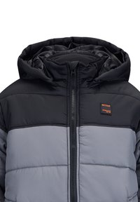 Gepolsterte Jacke mit einem schwarzen oberen Abschnitt und einem grauen unteren Abschnitt, mit einem Reißverschluss und einer Kapuze. Enthält ein auffälliges Logo-Patch.