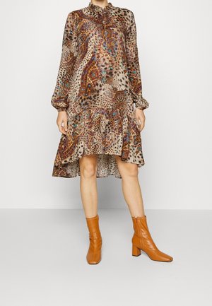 Model trägt ein gemustertes, langärmeliges Kleid mit einem rüschenbesetzten Saum und braunen Stiefeln mit Absatz und posiert vor einem schlichten weißen Hintergrund.