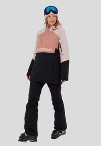 Vrouw in een roze en zwarte skijas, zwarte skibroek, zwarte skischoenen, roze beanie en skibril op het voorhoofd, staande voor een grijze achtergrond.