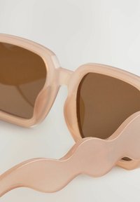 Vaaleanpunaiset aurinkolasit ruskeilla linsseillä, joissa on ainutlaatuinen aaltoileva muotoilu varsissa, valmistettu läpinäkyvästä muovimateriaalista.