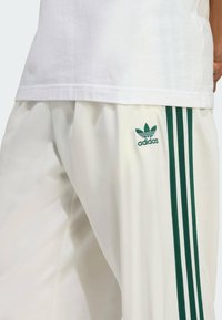 Vita joggerbyxor med tre gröna ränder längs sidorna och en broderad grön Adidas-logotyp på vänster lår, slät textur.