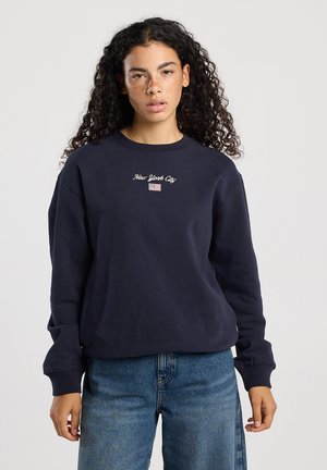 Giovane donna con capelli ricci indossa un felpa blu navy con ricamo "New York City" e bandiera degli Stati Uniti, abbinata a jeans blu, in piedi contro uno sfondo neutro.