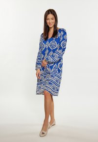 Rochie albastră cu motive ornamentale albe, mâneci lungi, decolteu în V și hem până la genunchi. Poartă pantofi plata bej. Design simplu, elegant.