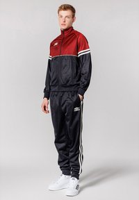 Träningsdräkt i röd och marinblå, med vita ränder, jacka med dragkedja och byxor med elastisk midja. Matchas med vita sneakers.