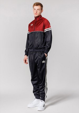 Trainingspak in rood en marineblauw, met witte strepen, een zippered jacket en broek met elastische tailleband. Gecombineerd met witte sneakers.