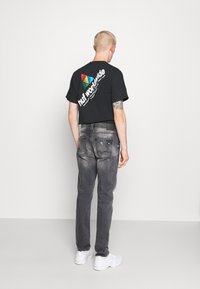 Camiseta negra de manga corta con un logotipo colorido en la parte posterior, combinada con jeans gris desgastados y zapatillas blancas, vista desde la parte de atrás.